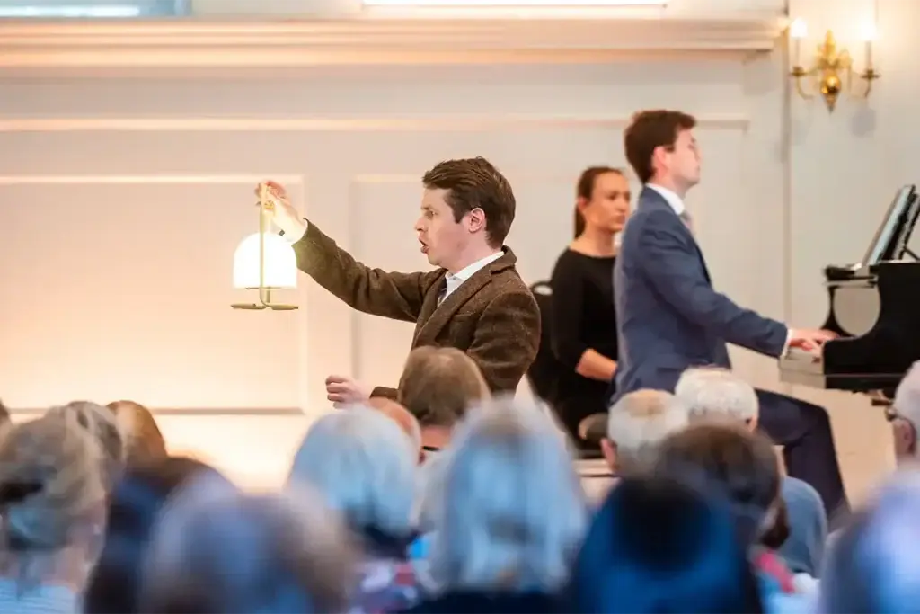 Man achtger een vleugel en een man met een lantaarn in een volle concertzaal.